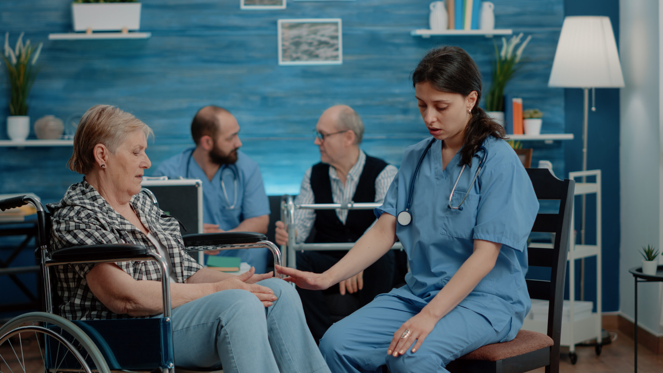 Nurse consulting retired woman sitting in wheelchair at nursing home clinic. Medical assistant doing healthcare checkup with disabled old patient for treatment and recovery from pain.